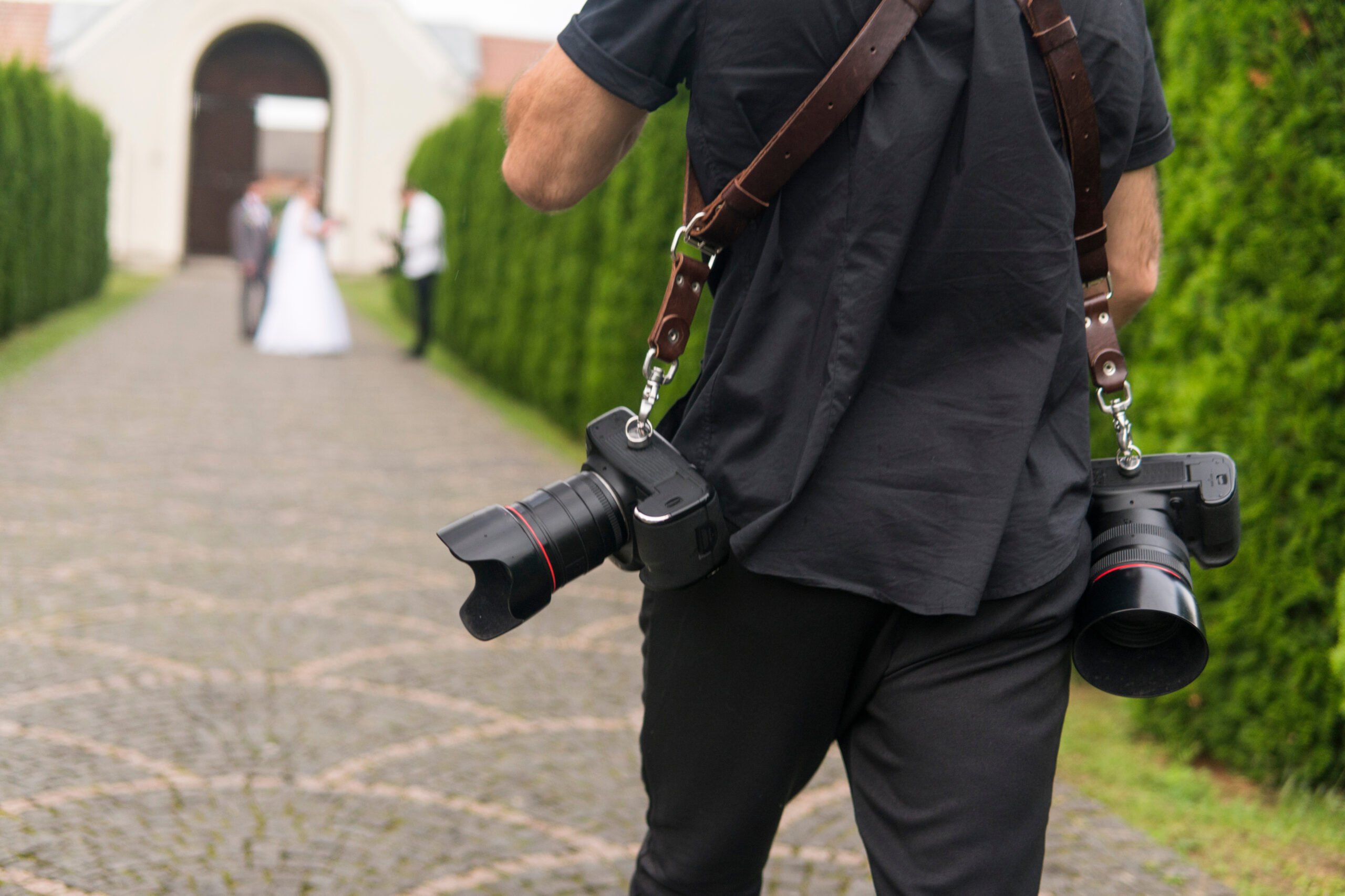 Professional wedding photographer with two cameras on a shoulder straps taking pictures of the bride and groom in a garden