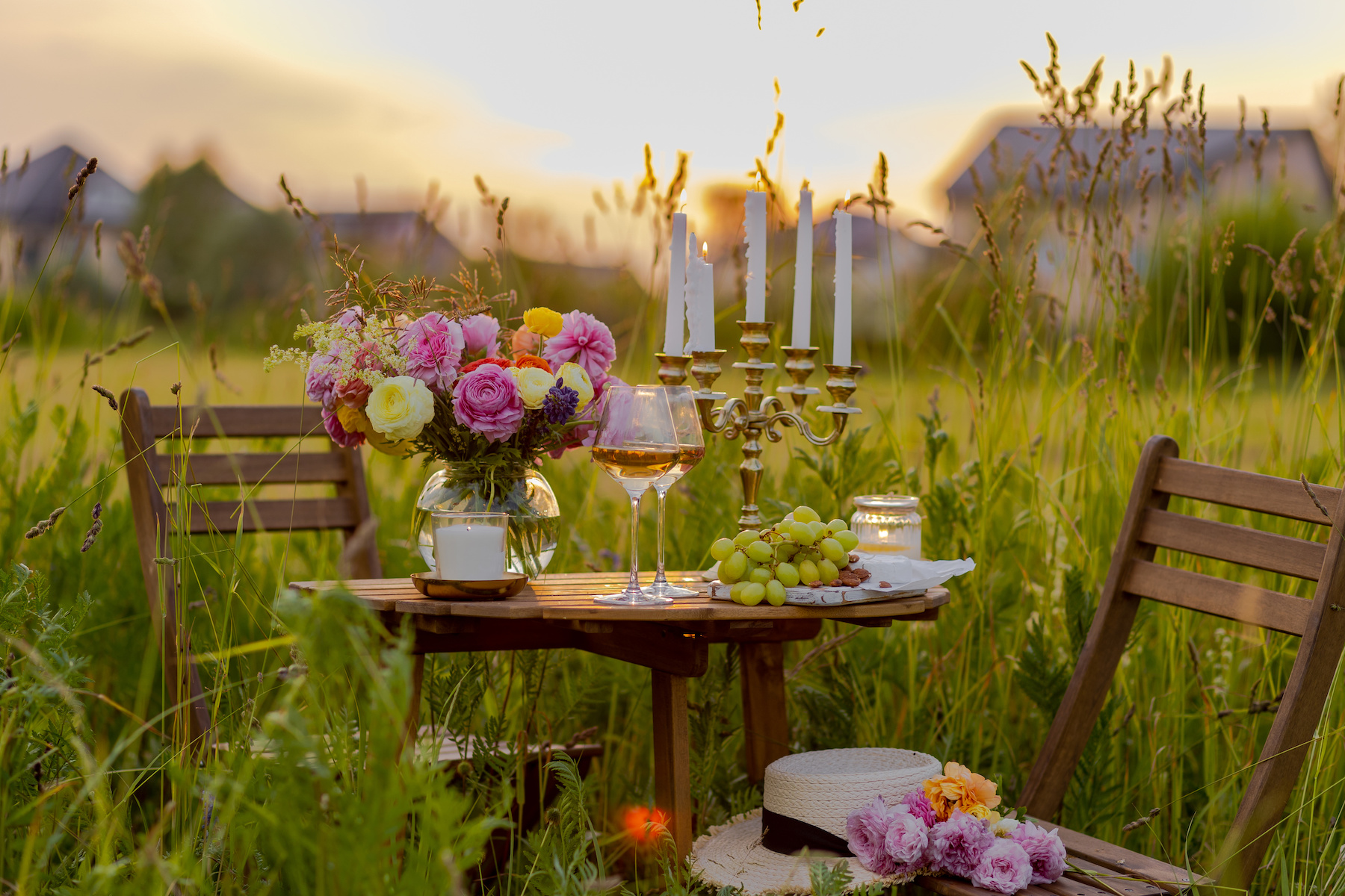 Romantic surprise, marriage proposal. Elegant decoration. Private wedding party for two, table set with floral decor, chandelier, fruits, wine in field or meadow. Summer, sunset, golden hour.